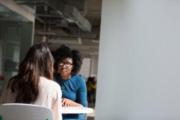 Woman at table speaking with another woman.