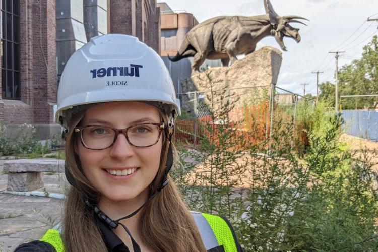 妮可Zalewski, wearing glasses and a construction helmet, stands in front of an old brick building with a statue of a triceratops out front.